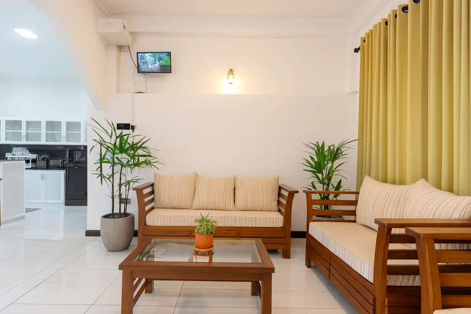 Katunayaka apartment sitting area with wooden sofas, cream cushions, plants and yellow curtains
